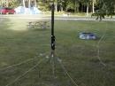 And finally the vertical.  Notice anything familiar in the picture -- yes it's my screwdriver antenna.  I set 9 radials at 20 degree increments starting at the NE and moving clockwise around the compass.  The picture shows the 8 ft whip attached as I had been checking 10m propagation.  The white wire hanging to the right is a 45ft wire radiator that replaced the whip for operation on 80m.  At first I could not find any resonance but after much checking I discovered that OHMIC connection of the radials to the base of the antenna is a requirement!  With the 8ft whip I normally have to load the coil almost all the way out for 80m.  With the 45ft radiator I only had to give it about an inch of coil -- that has to make a big improvement in radiation resistance.